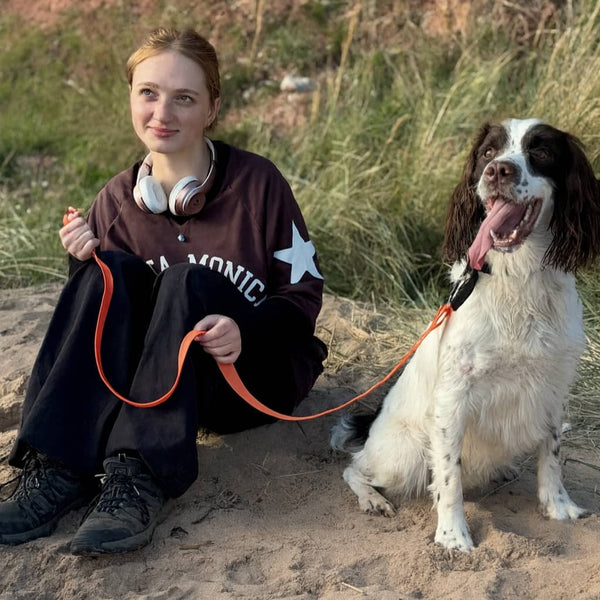 Woman sitting on beach with orange waterproof and non smelly lead attached to happy springer spaniel beside her on sand.